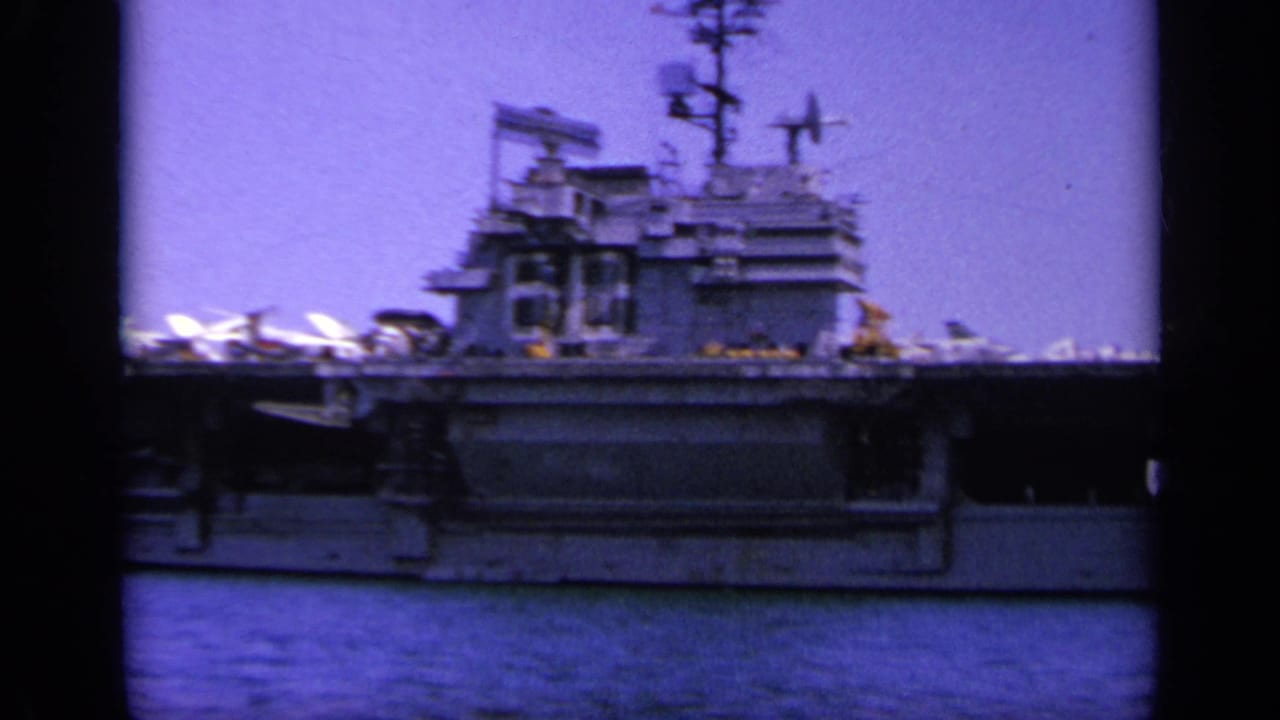 The carrier's island superstructure and deck aircraft fill the frame, filmed from a nearby vessel in 1963 Greece.