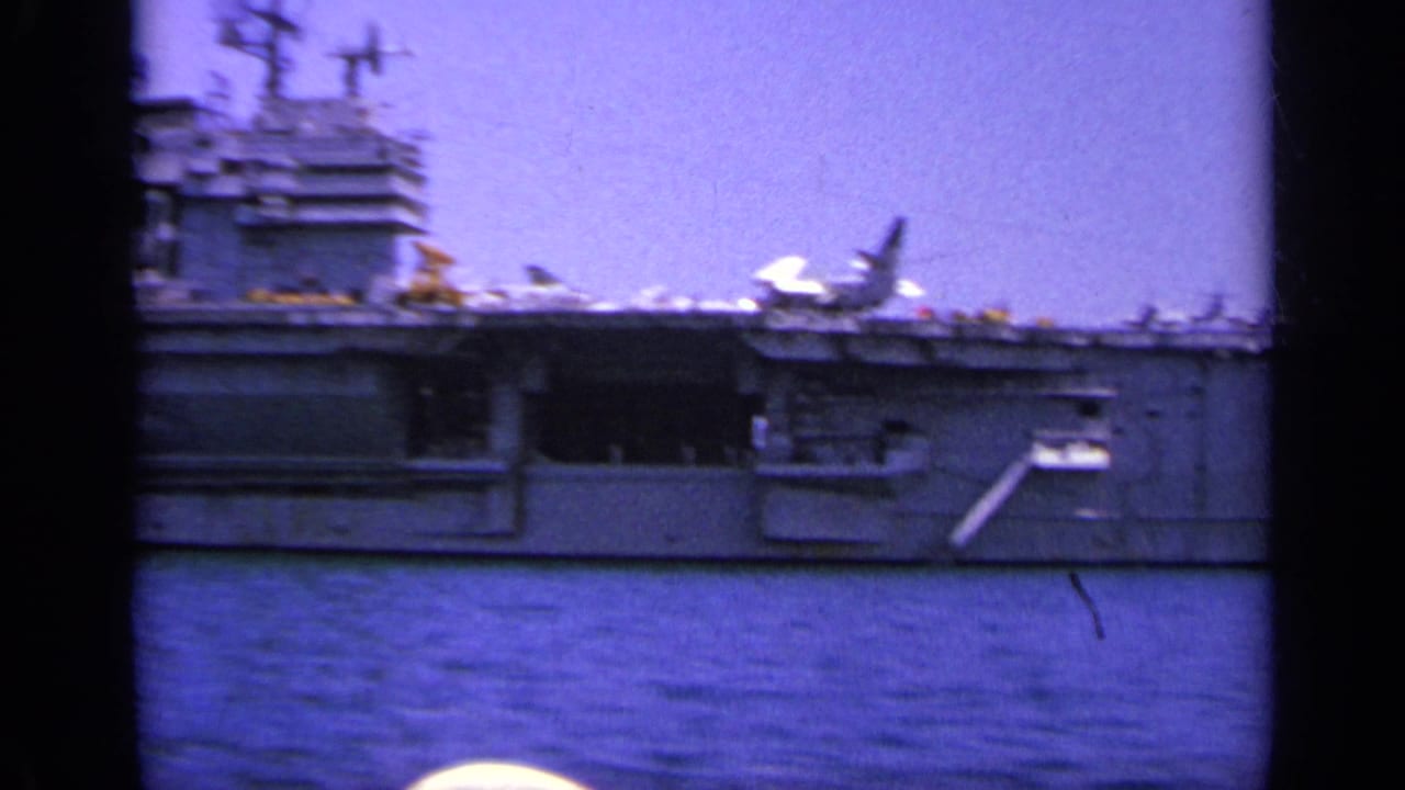 A handheld 8mm frame shows the carrier broadside, with a jet on deck and sailors or figures faintly visible along the rail.