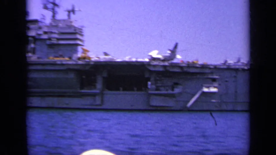 A handheld 8mm frame shows the carrier broadside, with a jet on deck and sailors or figures faintly visible along the rail.
