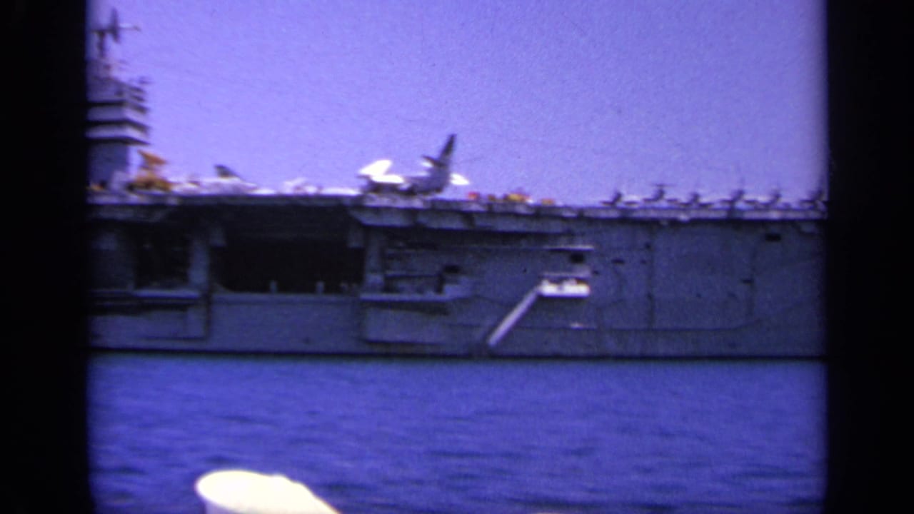 Aircraft carrier passing at sea with sailors' white caps in the foreground.