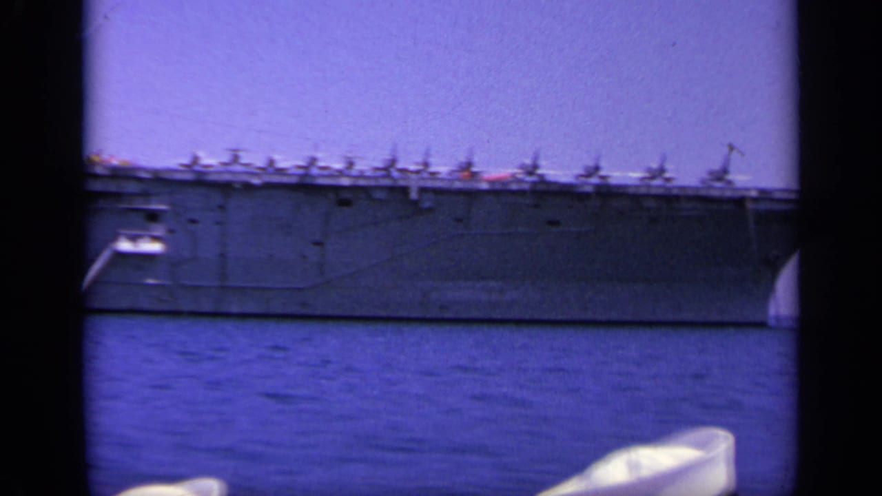 1963 Greece home-movie frame with a long side view of a gray aircraft carrier underway, deck aircraft visible against a clear sky.