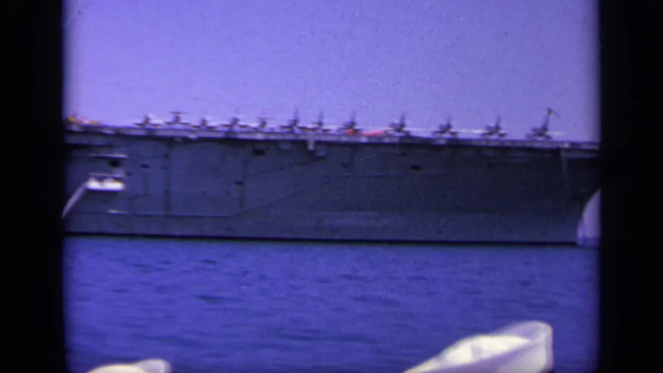 1963 Greece home-movie frame with a long side view of a gray aircraft carrier underway, deck aircraft visible against a clear sky.