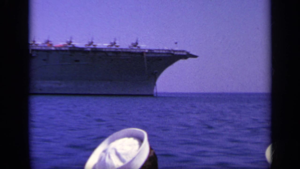 1963 Greece 8mm frame showing the bow of an aircraft carrier at sea, with white-capped sailors partly visible in the foreground.