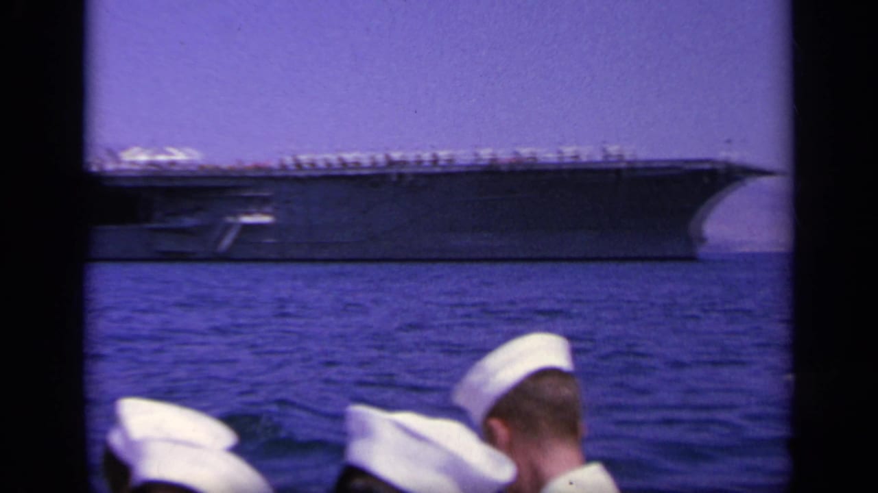 1963 Greece home-movie frame showing an aircraft carrier at sea beyond several sailors in white caps on the filming vessel.
