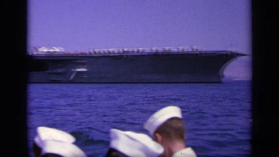 1963 Greece home-movie frame showing an aircraft carrier at sea beyond several sailors in white caps on the filming vessel.
