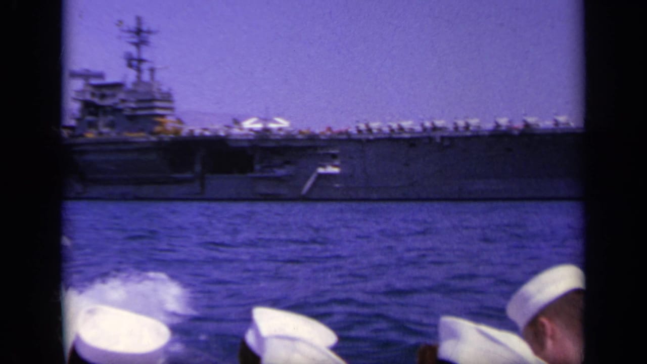 Aircraft carrier at sea seen past sailors in white caps on another vessel.