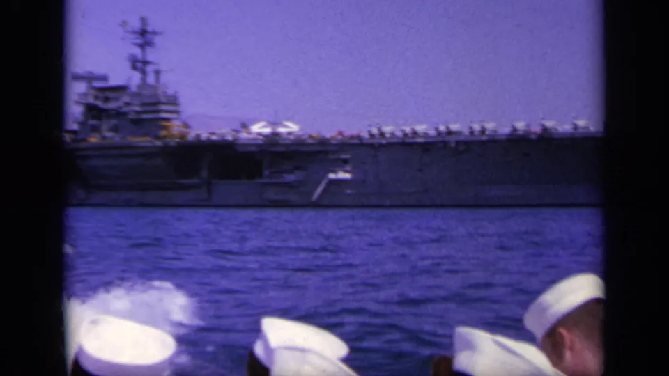 1963 8mm view of a U.S. Navy aircraft carrier at sea, filmed past sailors in white caps from another vessel.