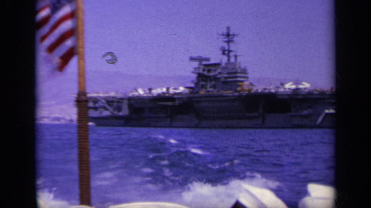 U.S. flag and wake in the foreground with an aircraft carrier beyond.