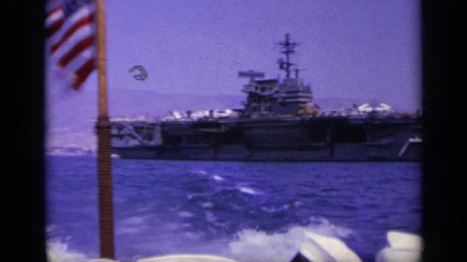 A U.S. flag flutters near the camera as a Navy aircraft carrier sits across the water in the 1963 home-movie frame.