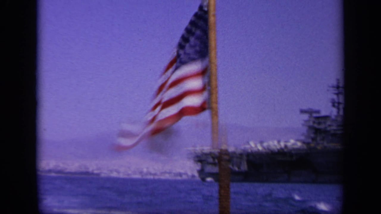 Handheld 8mm frame showing an American flag in the foreground and part of a U.S. Navy carrier across the water.