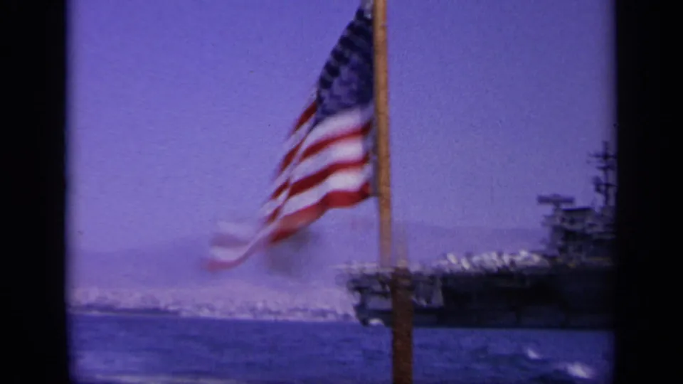 Handheld 8mm frame showing an American flag in the foreground and part of a U.S. Navy carrier across the water.