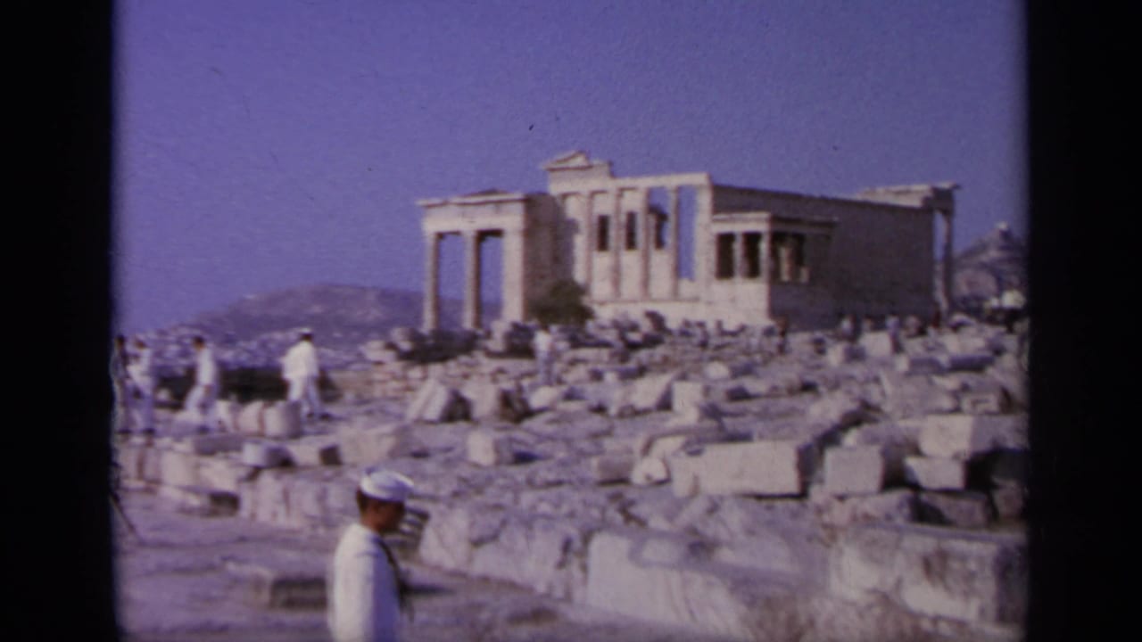 Sailors in white uniforms visit ancient ruins in Greece, with classical columns and scattered stone blocks visible.
