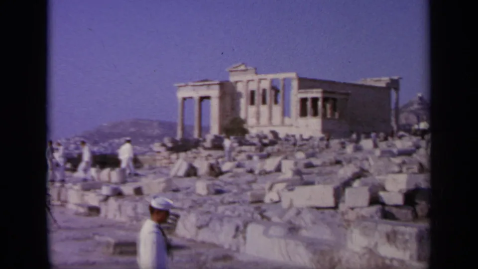 Sailors in white uniforms visit ancient ruins in Greece, with classical columns and scattered stone blocks visible.