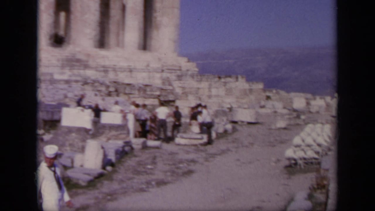 Tourists move along a pathway across the Acropolis ruins, with broken stonework and partial Parthenon columns visible nearby.