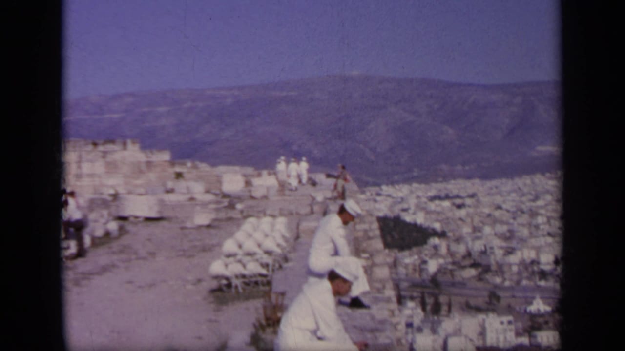 1963 Greece 8mm frame of two men in white uniforms at ancient stone ruins above a sprawling city and mountains.