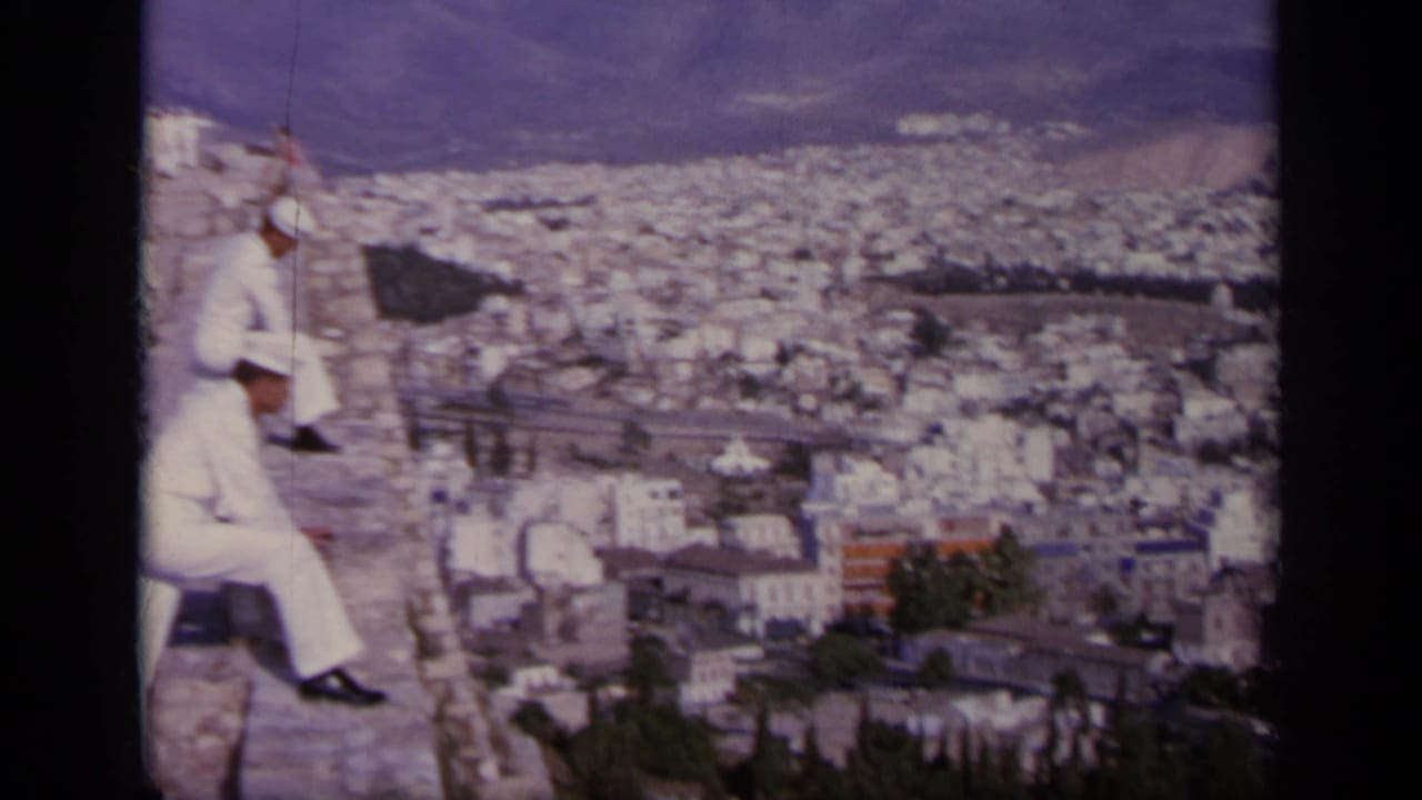1963 Greece home-movie frame showing uniformed men seated on ruins with a wide urban panorama in the background.