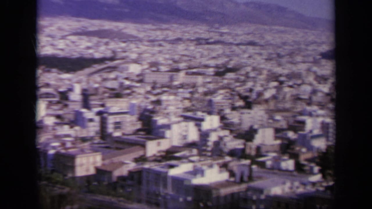 1963 Greece home-movie frame showing buildings, trees, and distant hills from a high overlook near ancient ruins.