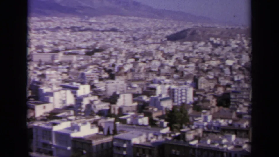 1963 8mm view over a dense Greek cityscape of pale buildings, flat roofs, scattered trees, and distant hills.