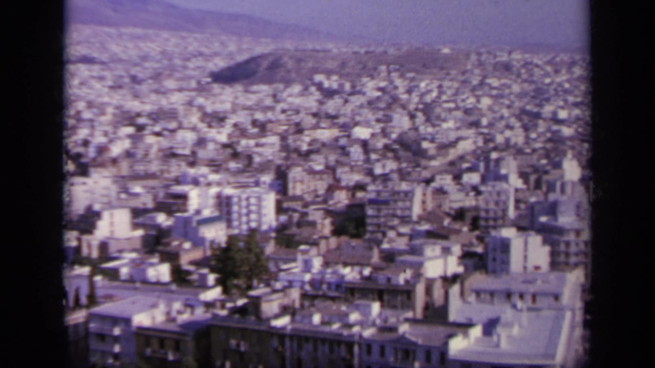 Urban panorama with pale buildings, rooftops, trees, and a low hill beyond the city.