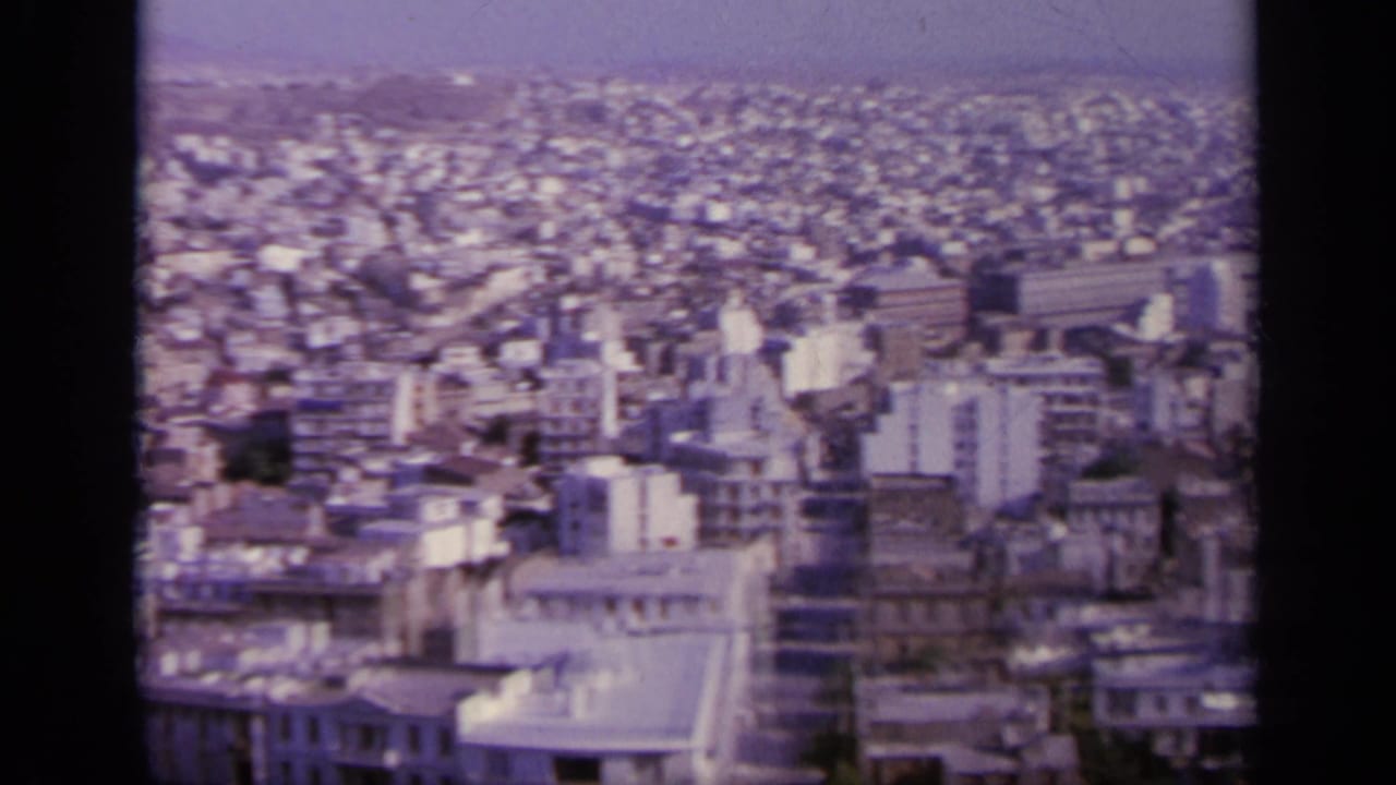 1963 8mm overlook showing a dense Greek urban area with apartment blocks, flat rooftops, streets, trees, and distant hills.