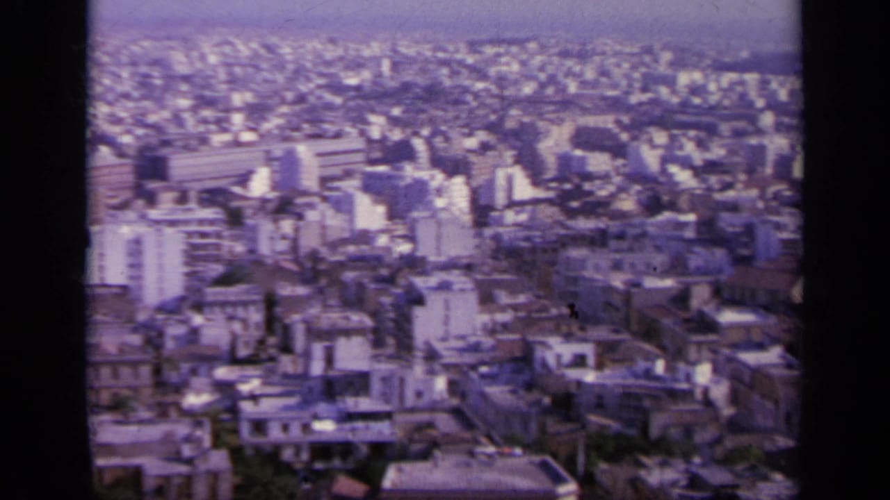 1963 Greek city view with a large rectangular building, dense rooftops, street lines, trees, and hills beyond the urban area.
