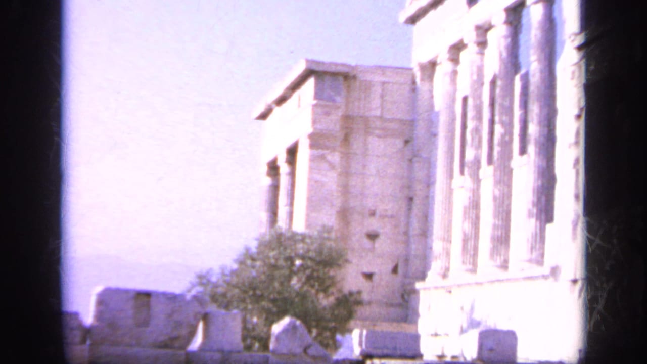 1963 8mm view in Greece with pale classical columns, a stone structure, a small tree, and overexposed sunlight.