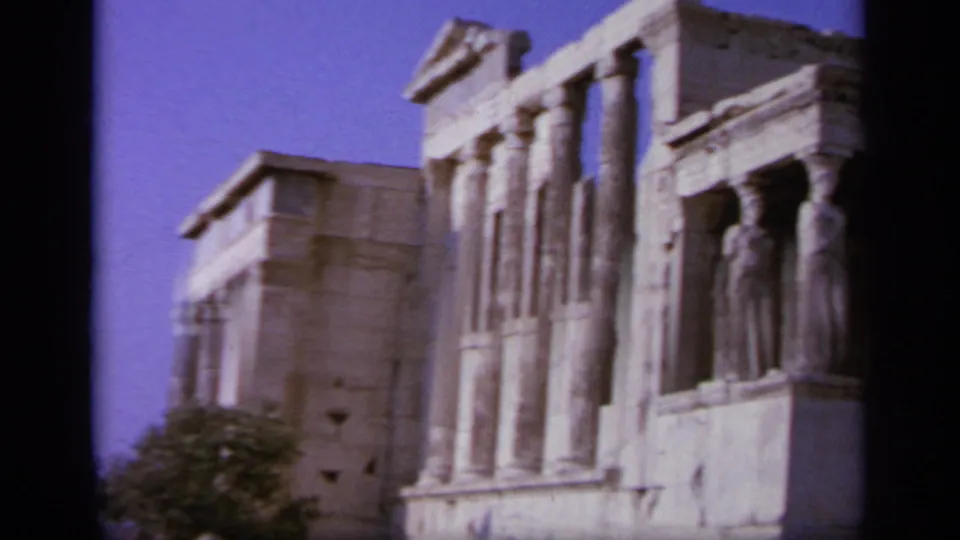 1963 Greece frame showing classical columns and carved standing figures on an ancient stone facade under a blue sky.