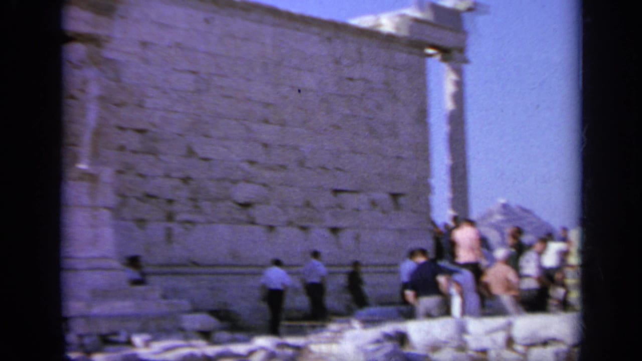 1963 Greece home-movie frame showing visitors in casual clothing near a large ancient stone wall and ruins in bright daylight.