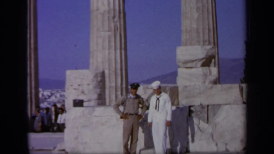 1963 Athens-area home-movie frame of a U.S. Navy sailor and tan-uniformed official posing among ancient marble columns.