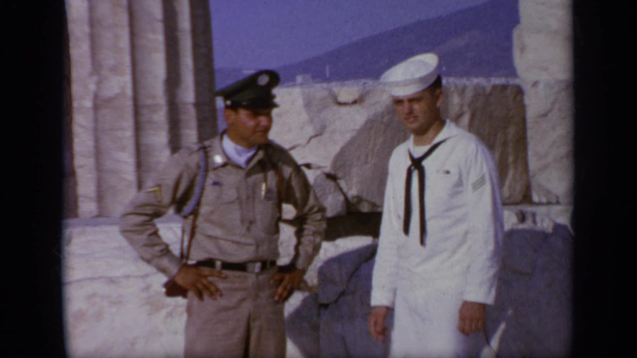 Closer 1963 home-movie frame from Greece showing the sailor and tan-uniformed official posed beside ancient stone blocks.