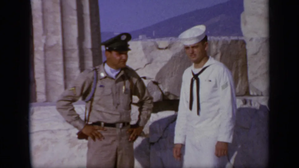 Closer 1963 home-movie frame from Greece showing the sailor and tan-uniformed official posed beside ancient stone blocks.