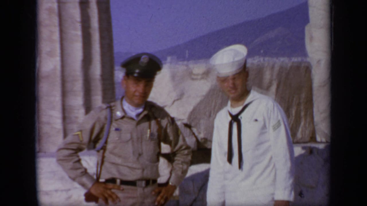 In Greece, 1963, a sailor in white and a tan-uniformed officer pose near ancient stone columns under a clear sky.