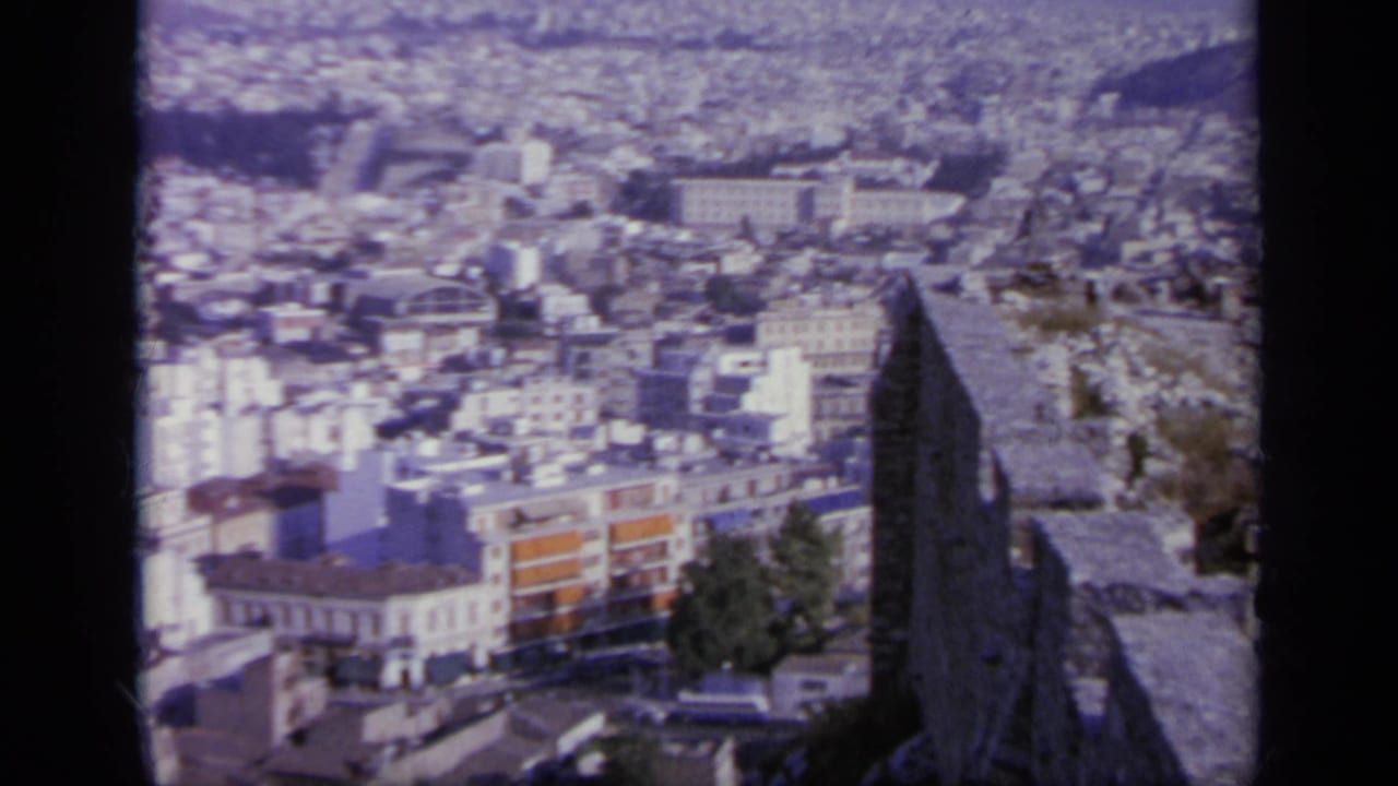 From an elevated ruin or terrace in Greece, the camera looks across dense white buildings and hills in soft 1960s color.