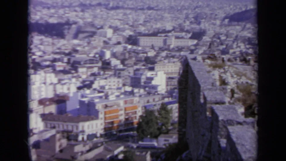 From an elevated ruin or terrace in Greece, the camera looks across dense white buildings and hills in soft 1960s color.
