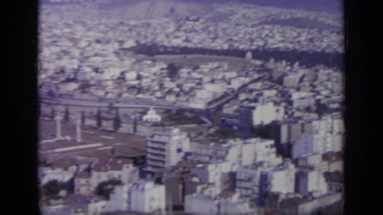 1963 Greece home-movie frame showing a high-angle city view with pale buildings, ancient ruins, and hills beyond.