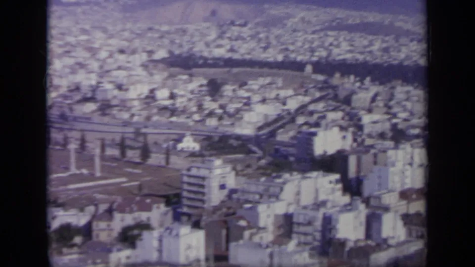 1963 Greece home-movie frame showing a high-angle city view with pale buildings, ancient ruins, and hills beyond.