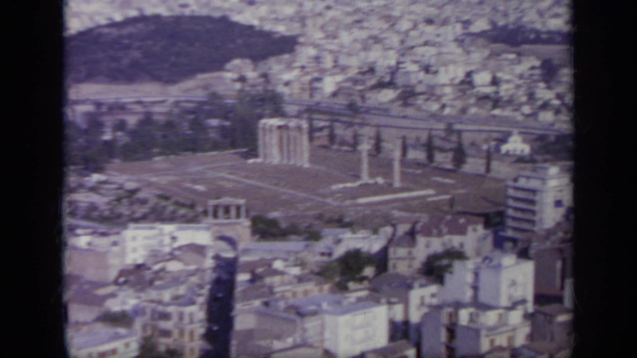 High view of city blocks, ancient columns, roads, and a wooded hill.