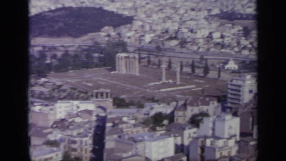 1963 Greece footage frame with ancient columns, roads, clustered buildings, and a dark hill behind the city.