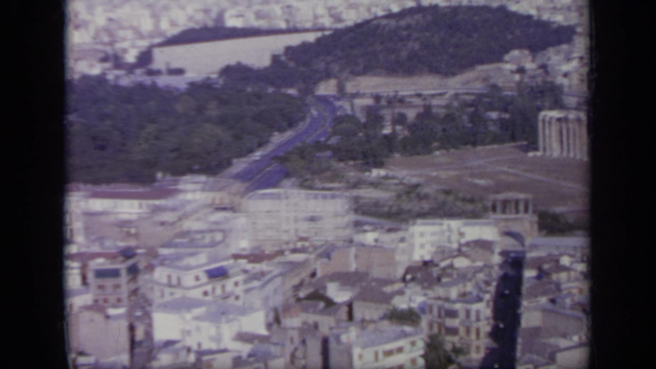 1963 Greece home-movie view over roads, ruins, trees, and dense buildings, with a hazy distant area beyond.