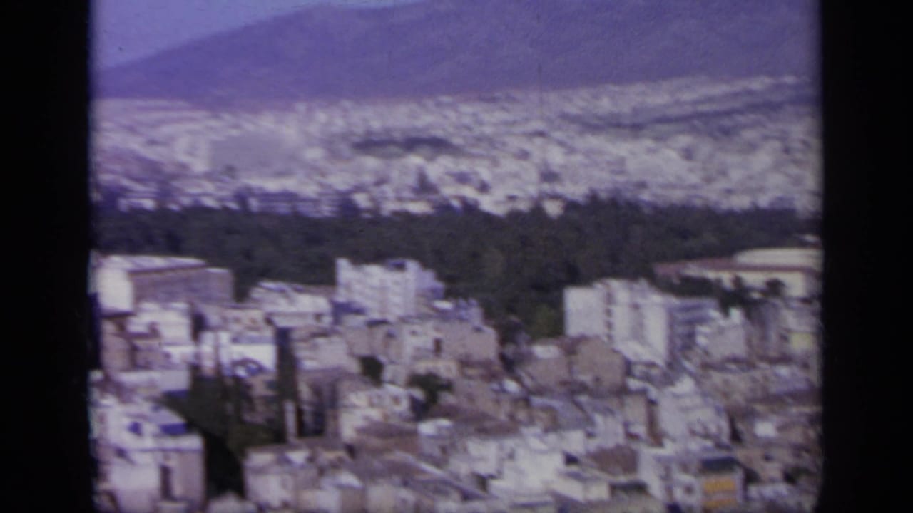 High view over a dense Greek city with white buildings, dark trees, hazy hills, and sky.
