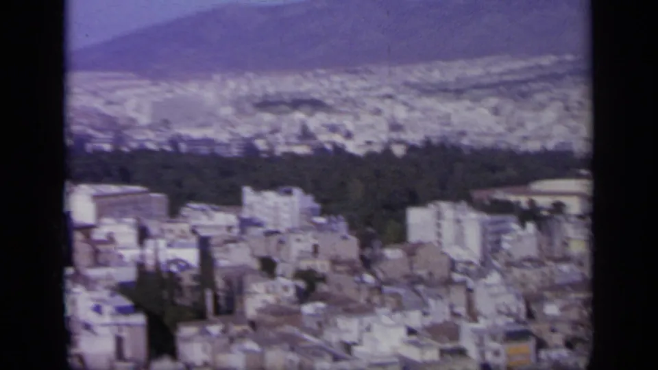 1963 8mm view over a Greek city in a valley, with pale buildings, a dark wooded band, and hazy hills beyond.
