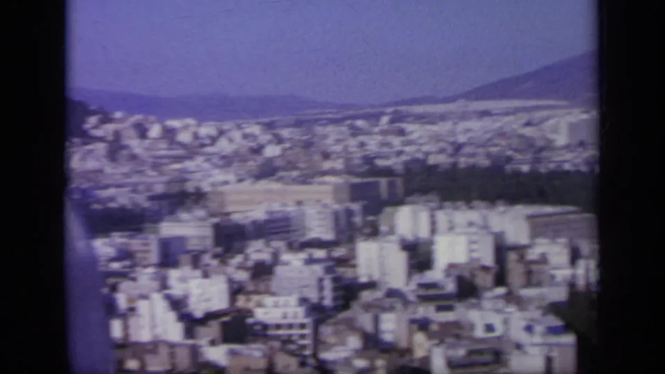 1963 Greek city panorama with white buildings, distant hills, and a dark mountain slope beyond the urban area.