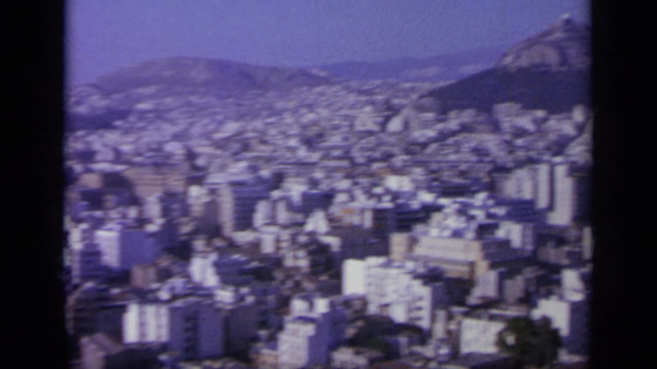 1963 8mm frame showing a Greek city below a rocky mountain slope, with dense buildings and haze across the valley.
