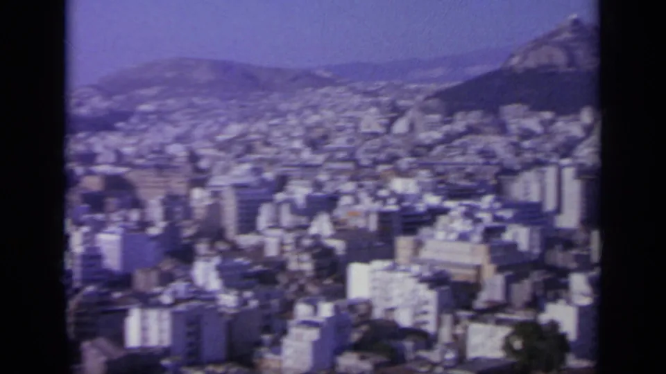 1963 8mm frame showing a Greek city below a rocky mountain slope, with dense buildings and haze across the valley.