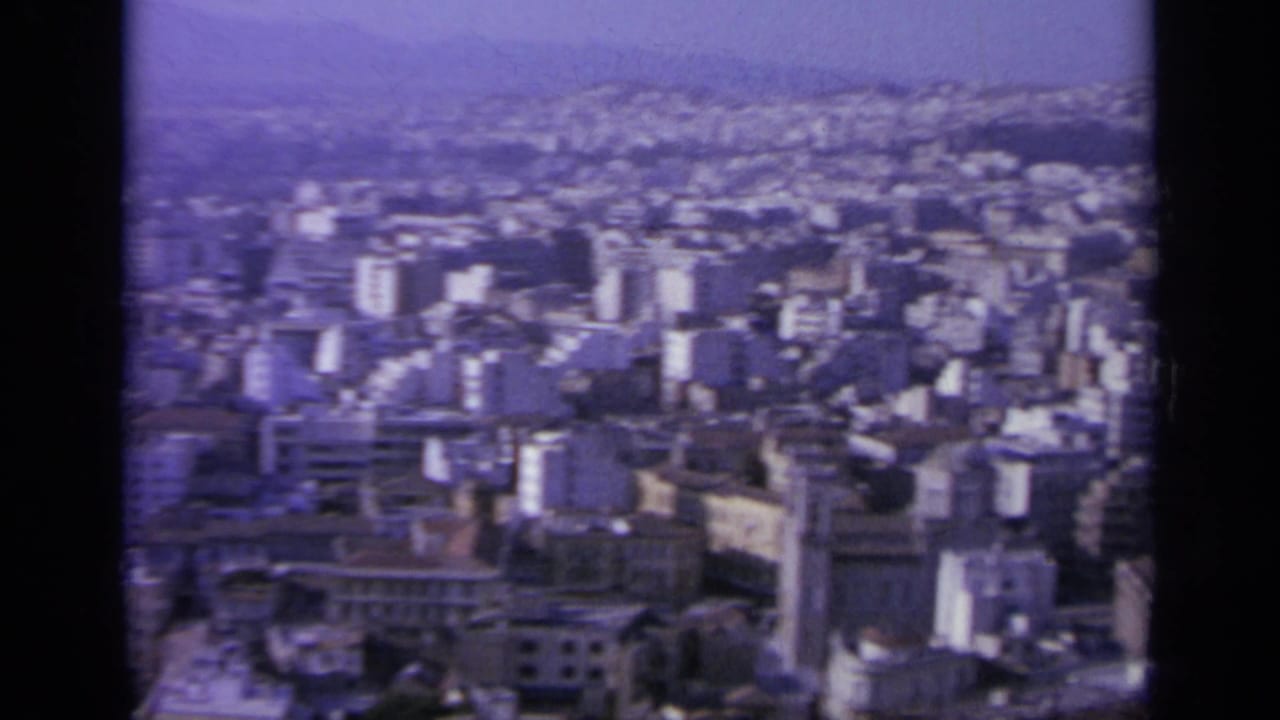 Wide 1963 Greek city view with mid-rise buildings in the foreground, dense hillside development, and distant hazy ridges.