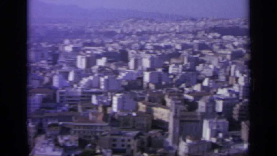 Wide 1963 Greek city view with mid-rise buildings in the foreground, dense hillside development, and distant hazy ridges.