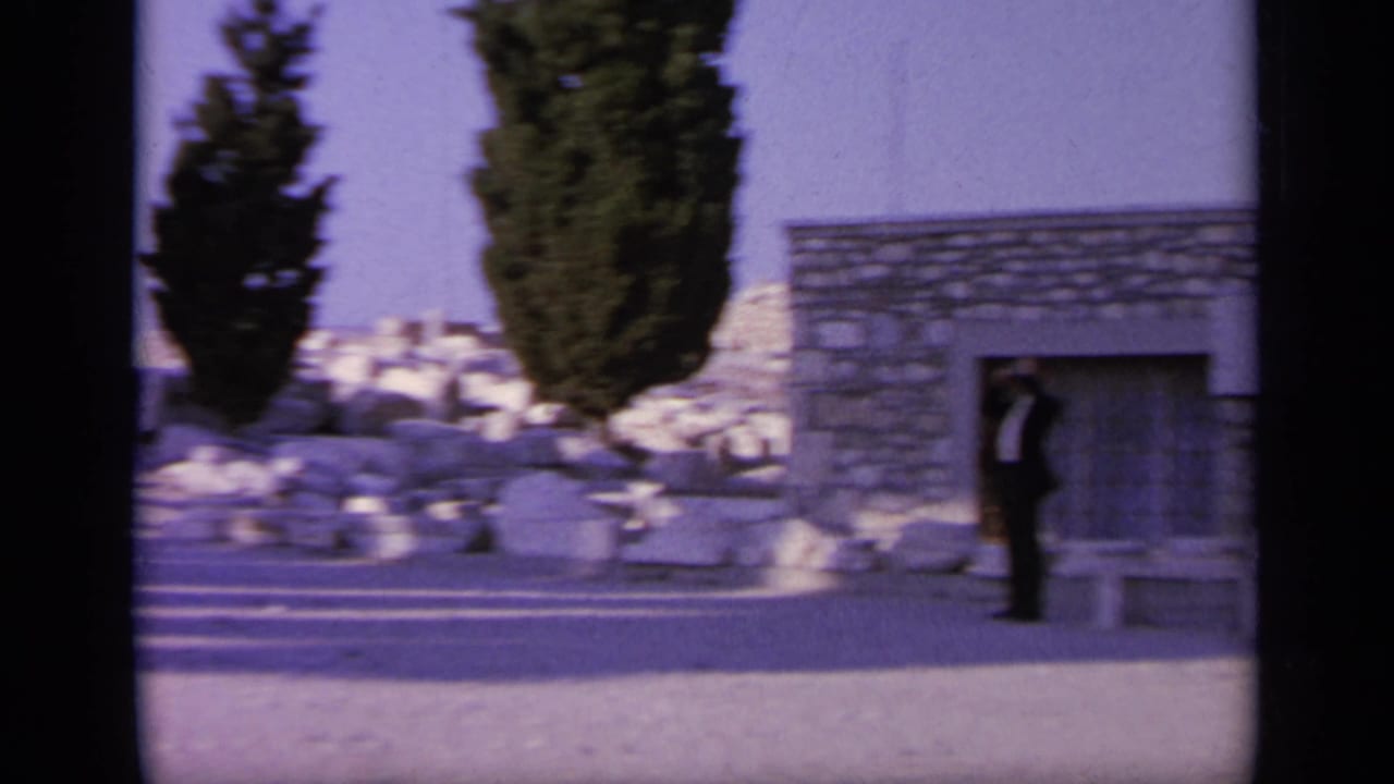 Man standing near a stone building with benches, trees, and ruins nearby.