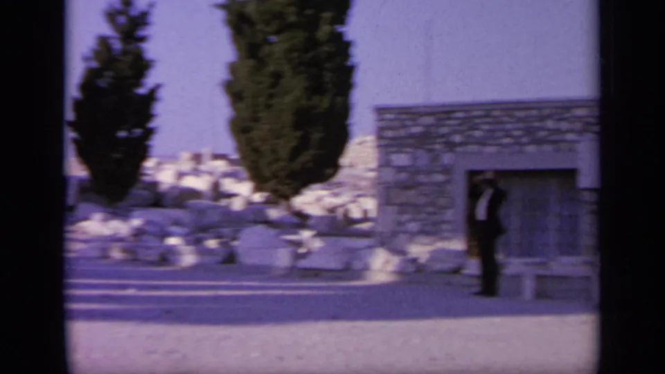 1963 Greece home-movie frame showing a man in dark clothing beside a low stone building, benches, trees, and scattered ruins.