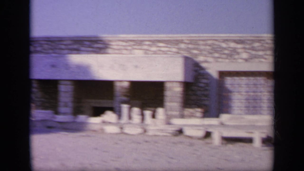 1963 Greece frame showing a low classical-style stone structure with columns, benches, and pale ruins under strong daylight.