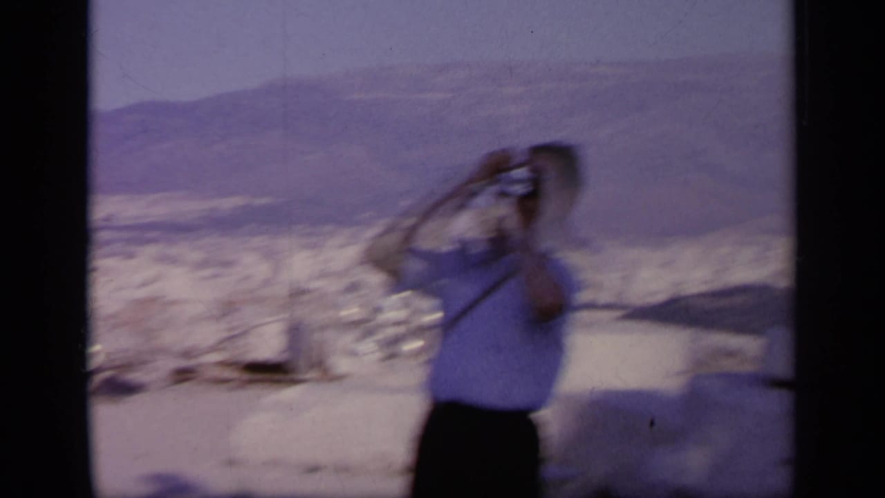 1963 Greece home-movie frame with a blurred tourist lifting a camera, backed by hills and pale stone ruins.